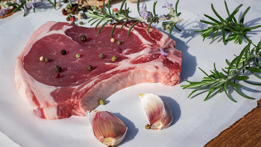 Belle côte de veau crue présentée avec quelques gousses d’ail et des feuilles de romarin, des billes de poivre.